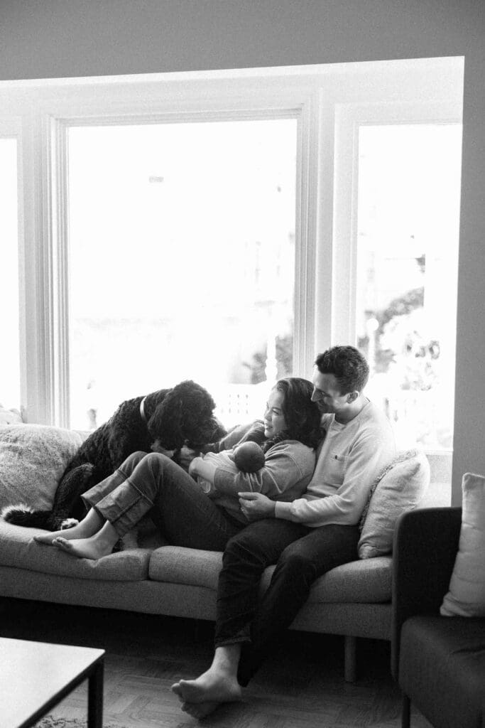New mom and dad cuddling on the couch with their new baby girl and their dog in front of a bay window in San Francisco