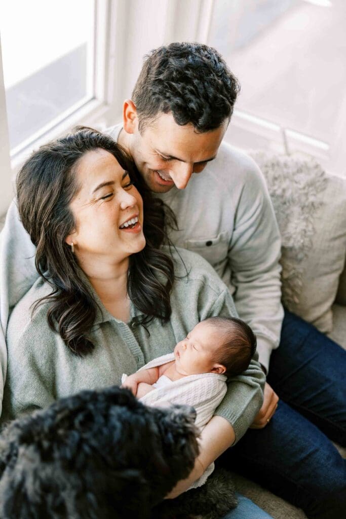 New mom and dad cuddling on the couch with their new baby girl and their dog in front of their bay window in San Francisco