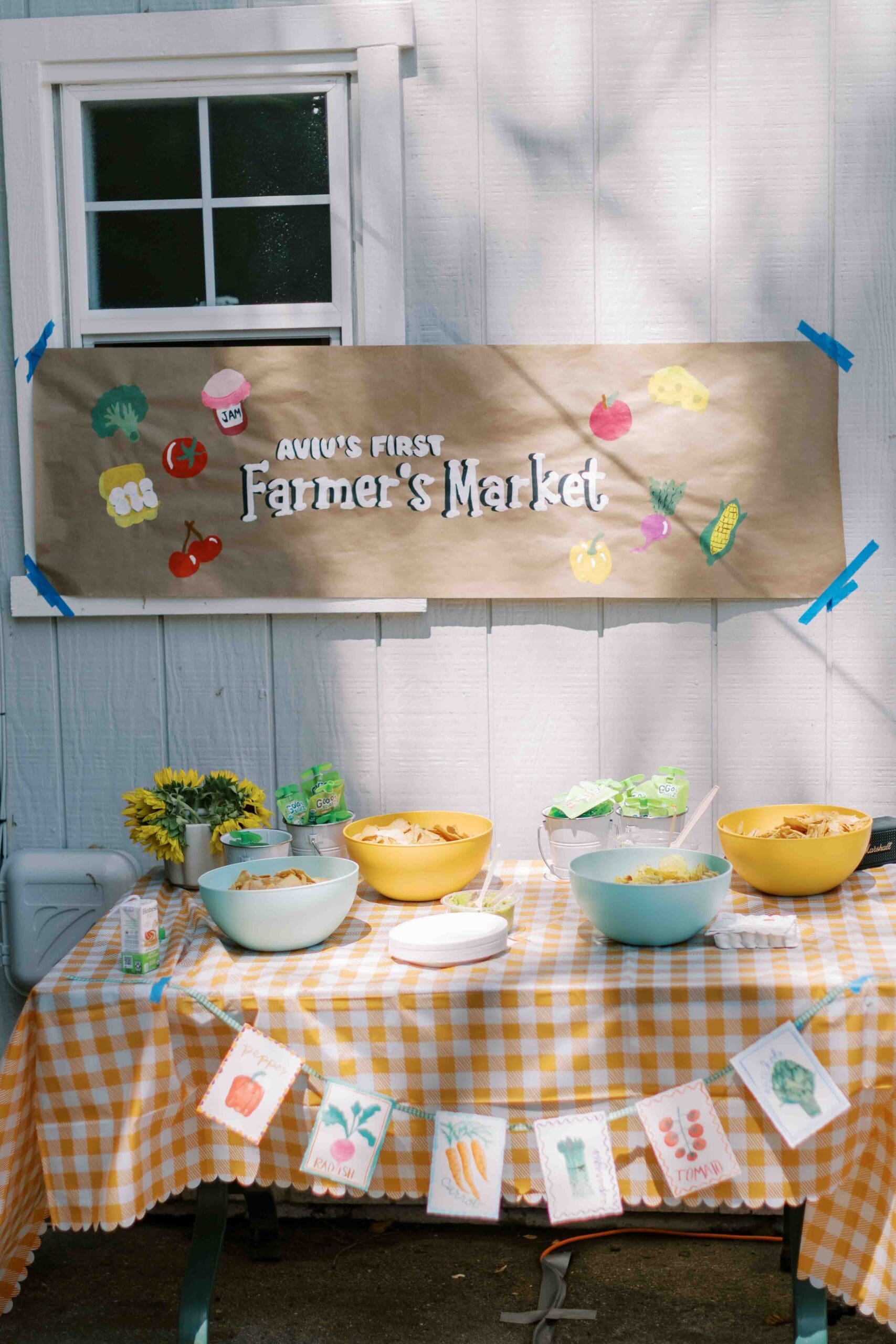 sign that says "aviv's first farmer's market" with a snack table covered in a yellow gingham tablecloth