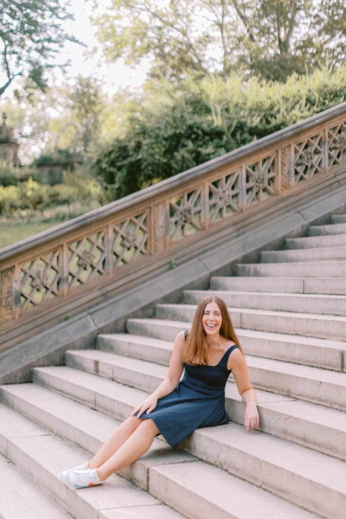 Woman wearing a navy blue skater dress and sitting on marble steps, smiling at the camera for her dating profile photos