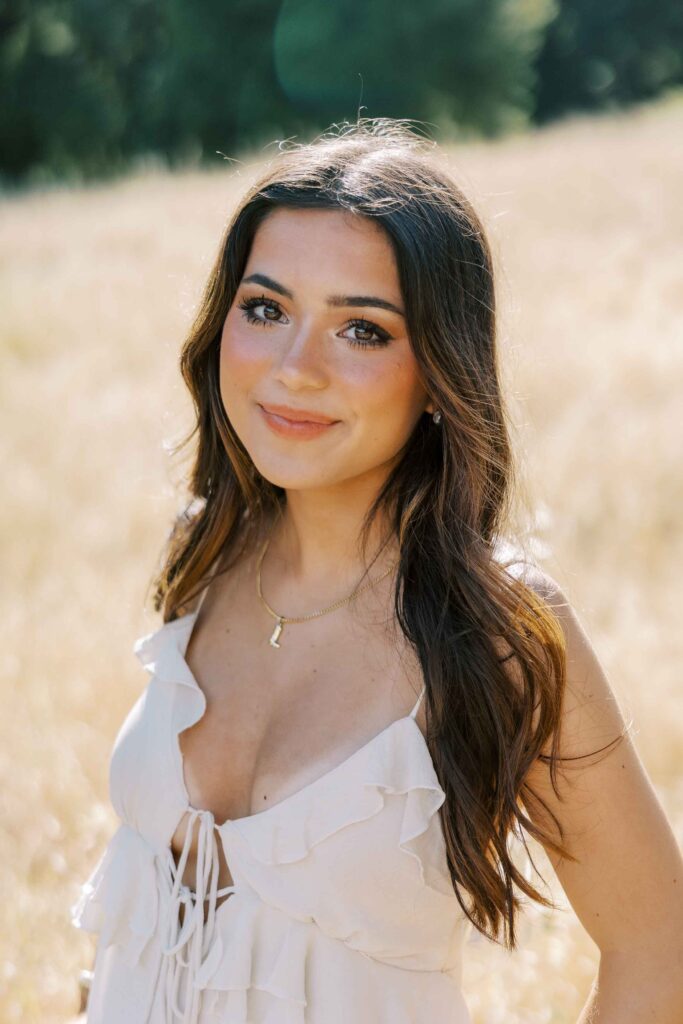 Brunette girl smiling with a soft smile at the camera, wearing a white dress and standing in an area with dry grass