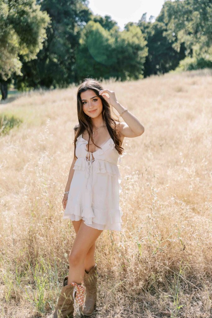 Brunette girl smiling with a soft smile at the camera, wearing a white dress and standing in an area with dry grass