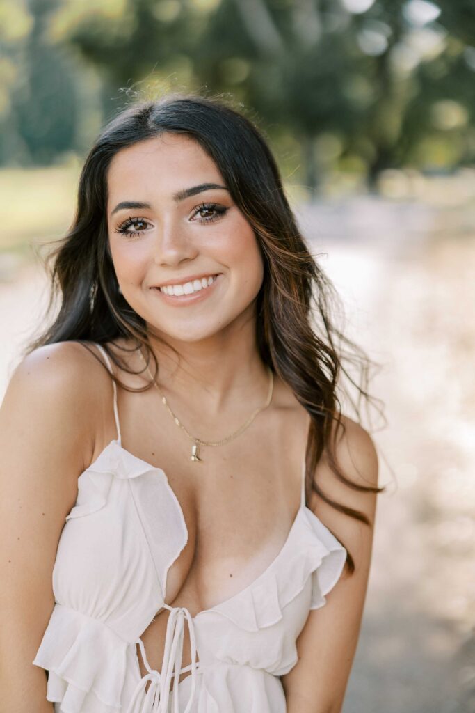 Brunette girl smiling with a big smile at the camera, wearing a white dress and standing in an area with dry grass
