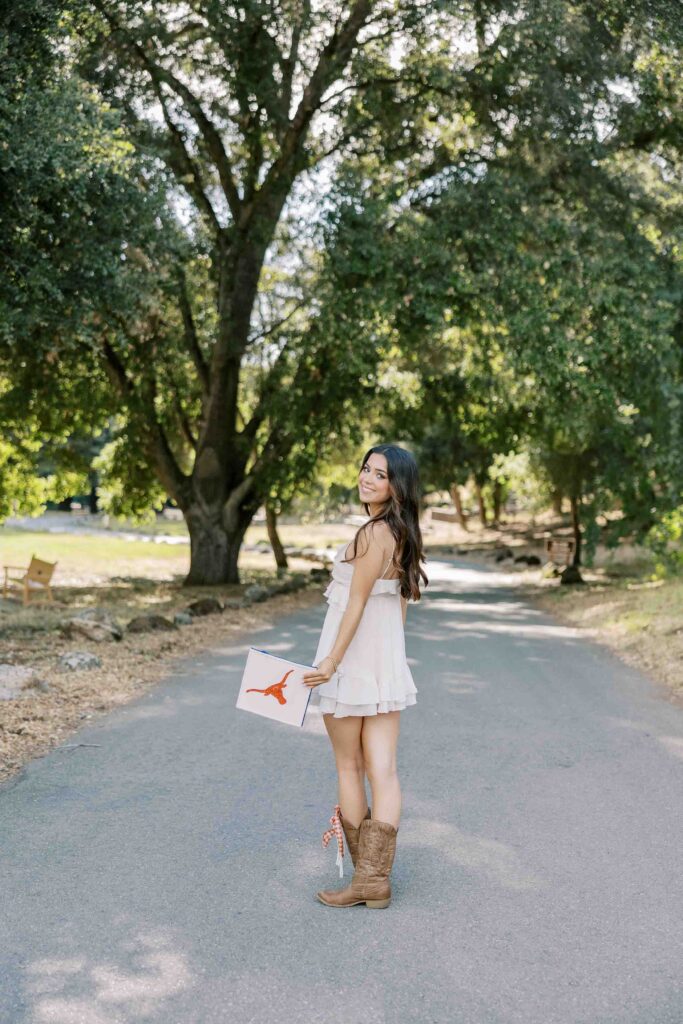 Brunette girl wearing a white dress and cowboy boots and looking back over her shoulder while holding a graduation cap