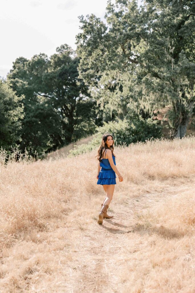 Brunette girl wearing a royal blue dress and looking over her shoulder while walking through tall, dry grass