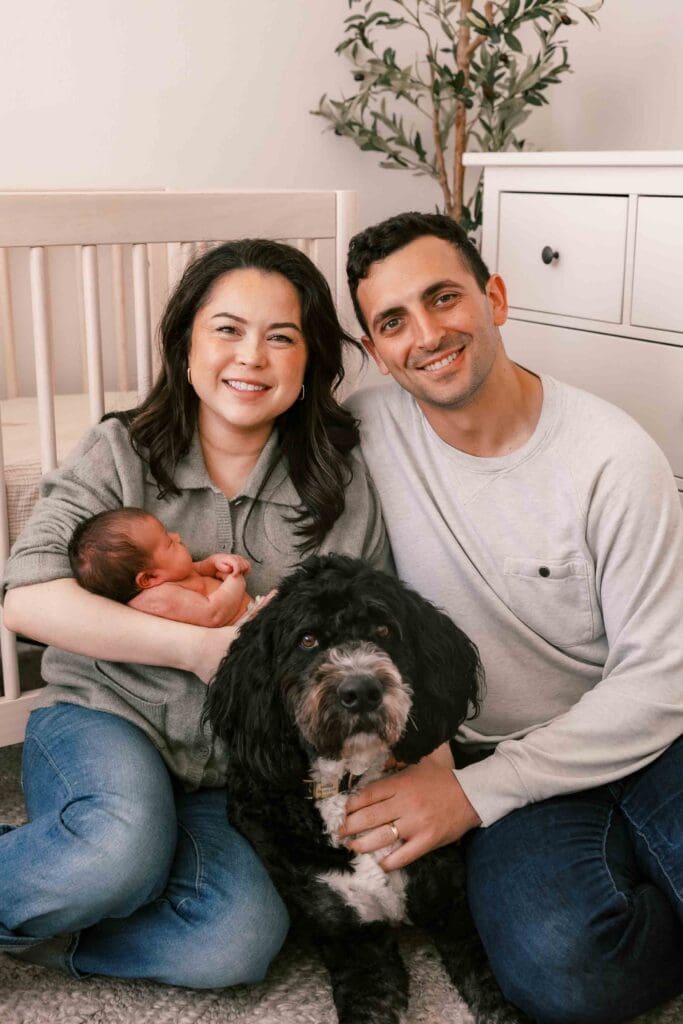New mom and dad holding their newborn baby with their bernedoodle looking at the camera