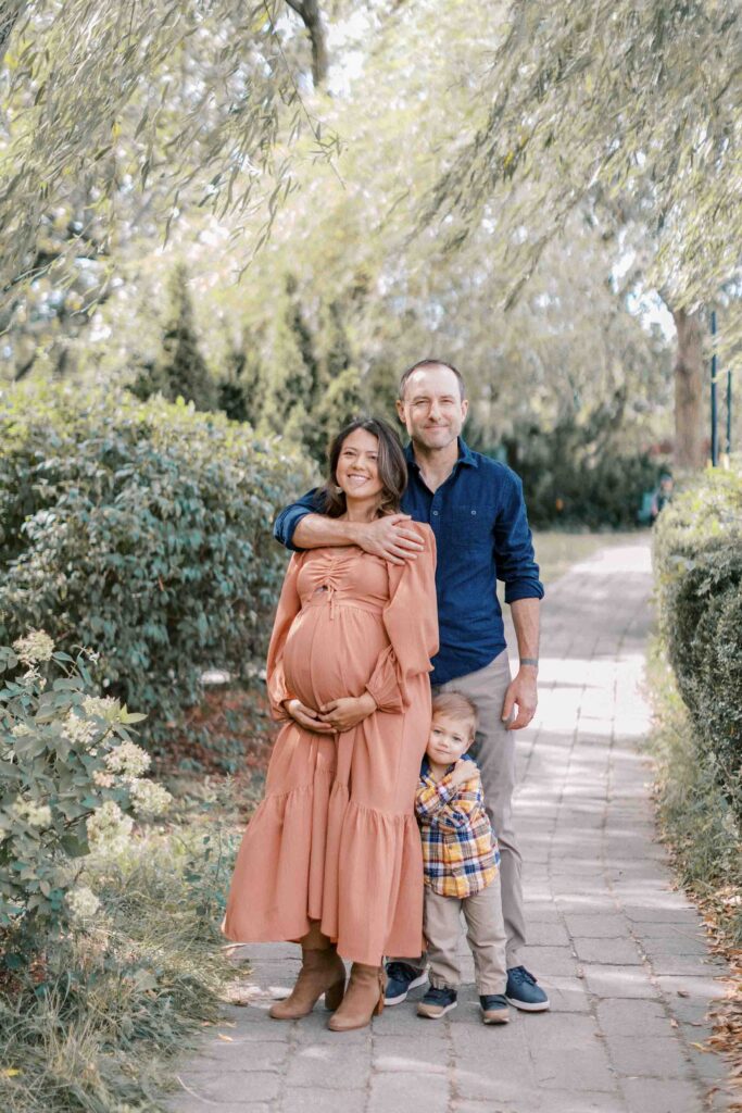 Maternity portrait of a mom, dad, and toddler boy standing in a green park