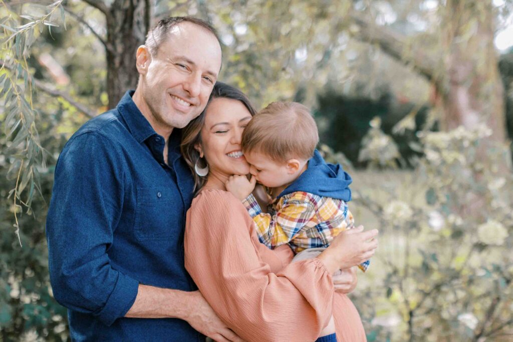 Maternity portrait of a mom, dad, and toddler boy standing in a green park and cuddling