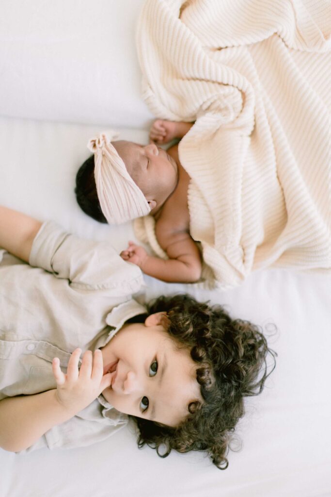 Toddler boy looking up at camera while laying next to his new baby sister