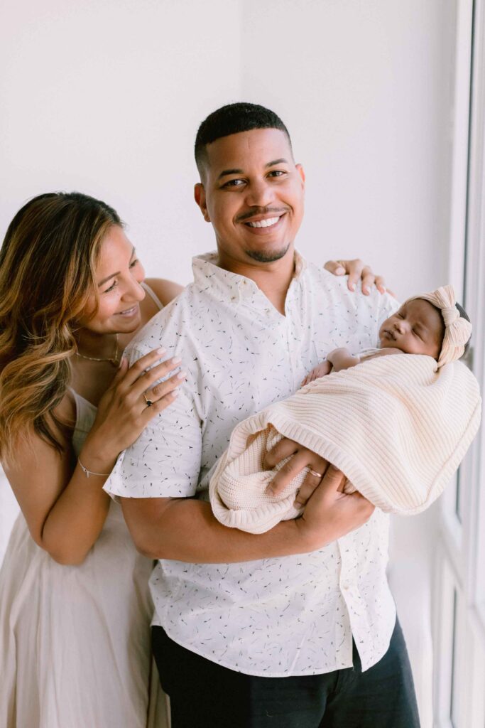 Mom and dad smiling and holding their new baby girl, wrapped in a cream colored blanket with a bow on her head