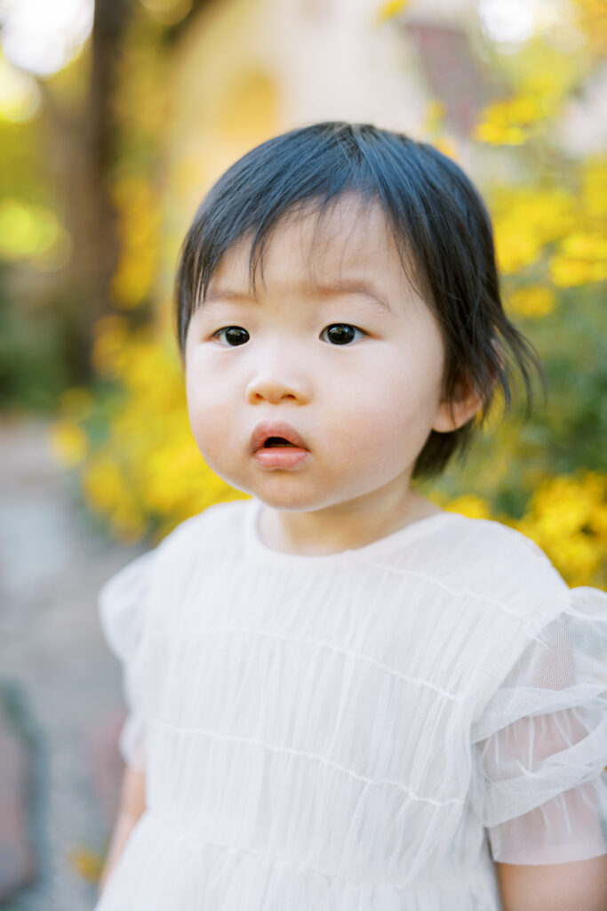 Toddler girl wearing a white dress, standing in front of yellow florals